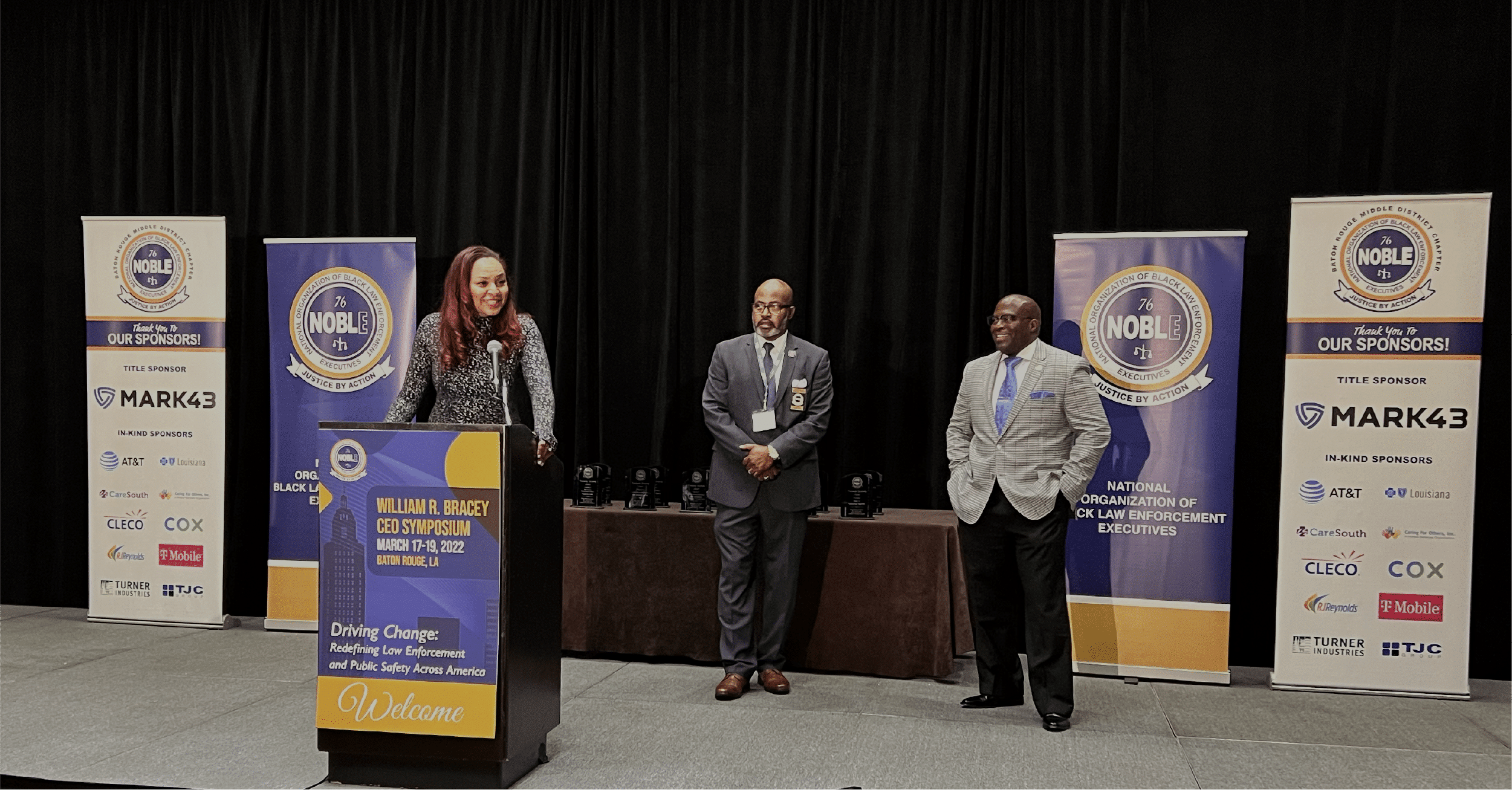Ganesha Martin, Mark43 VP of Community Affairs and Public Policy, is standing on a raised platform and behind a lectern while speaking at NOBLE. Two gentlemen are standing on the raised platform behind her.
